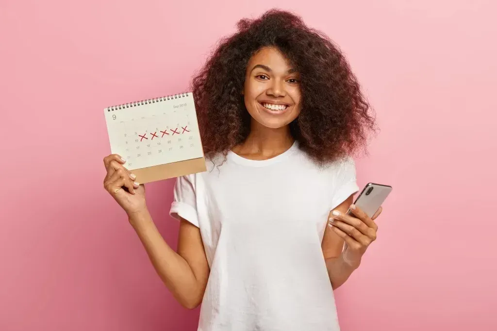 Black woman smiling, holding a calendar in one hand and a phone in the other — symbolizing the best menstrual cycle tracking apps of 2025 Belle Health