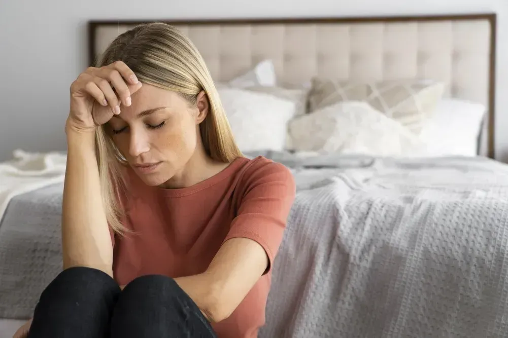 Frau sitzt nachdenklich auf dem Boden vor dem Bett und hält sich den Kopf – mögliches Symptom von Schwindel oder Kreislaufproblemen vor der Periode.