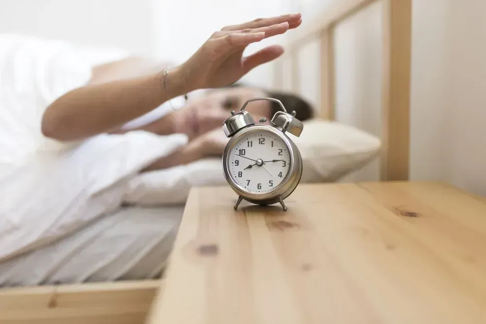 Frau liegt im Bett und greift nach dem Wecker auf dem Nachttisch – symbolisches Bild für ADHS und morgens früh aufstehen