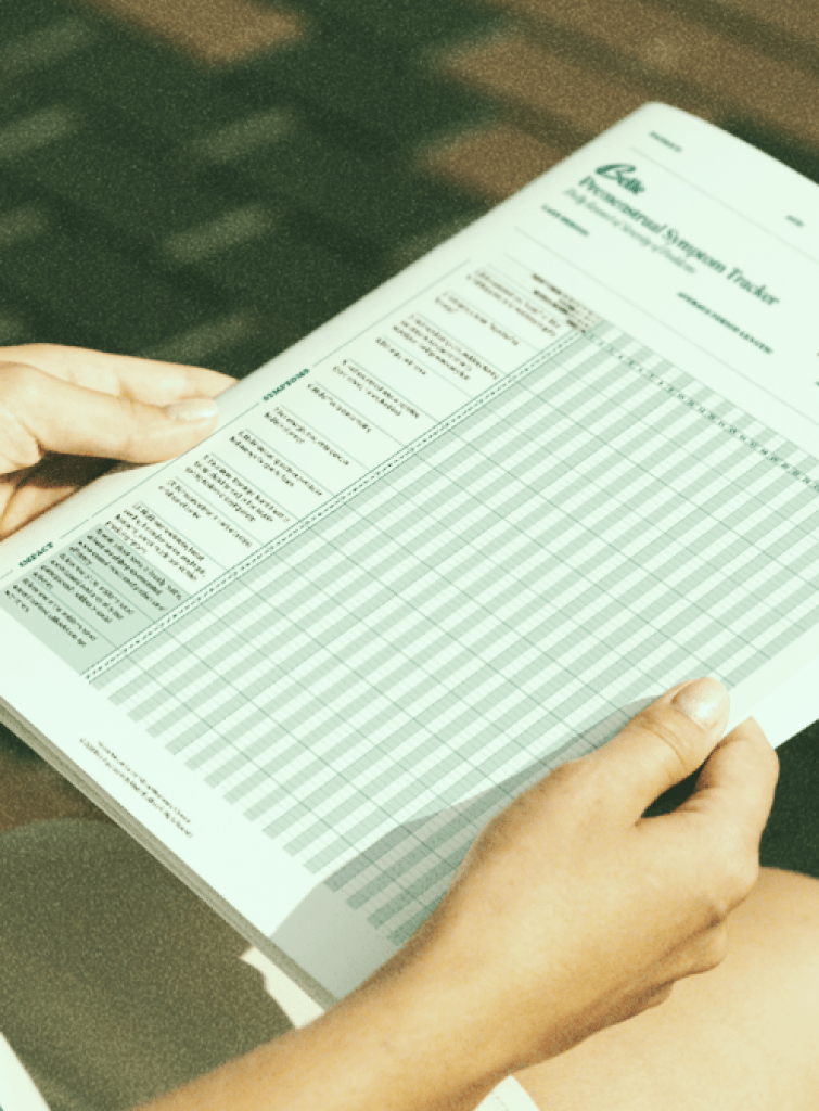 A person holding a printed "PMDD Symptom Tracker," which features a grid for tracking daily symptoms related to Premenstrual Dysphoric Disorder (PMDD). The chart includes rows for different symptoms, including physical, emotional, and behavioral aspects, and columns for each day of the month to help monitor symptom patterns over time.