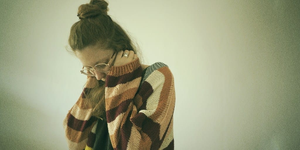 A young woman with red hair, wearing glasses and a striped knit sweater, showing anxiety during ovulation. She is holding her head in her hands, leaning slightly forward, with a pensive and overwhelmed expression against a muted, plain background.