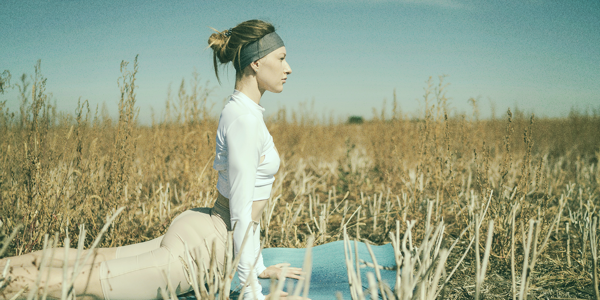 A woman practices yoga in a field, performing the upward-facing dog pose on a yoga mat. She is dressed in athletic wear, and the setting is a serene, dry landscape under a clear blue sky, representing how self-care can help PMDD