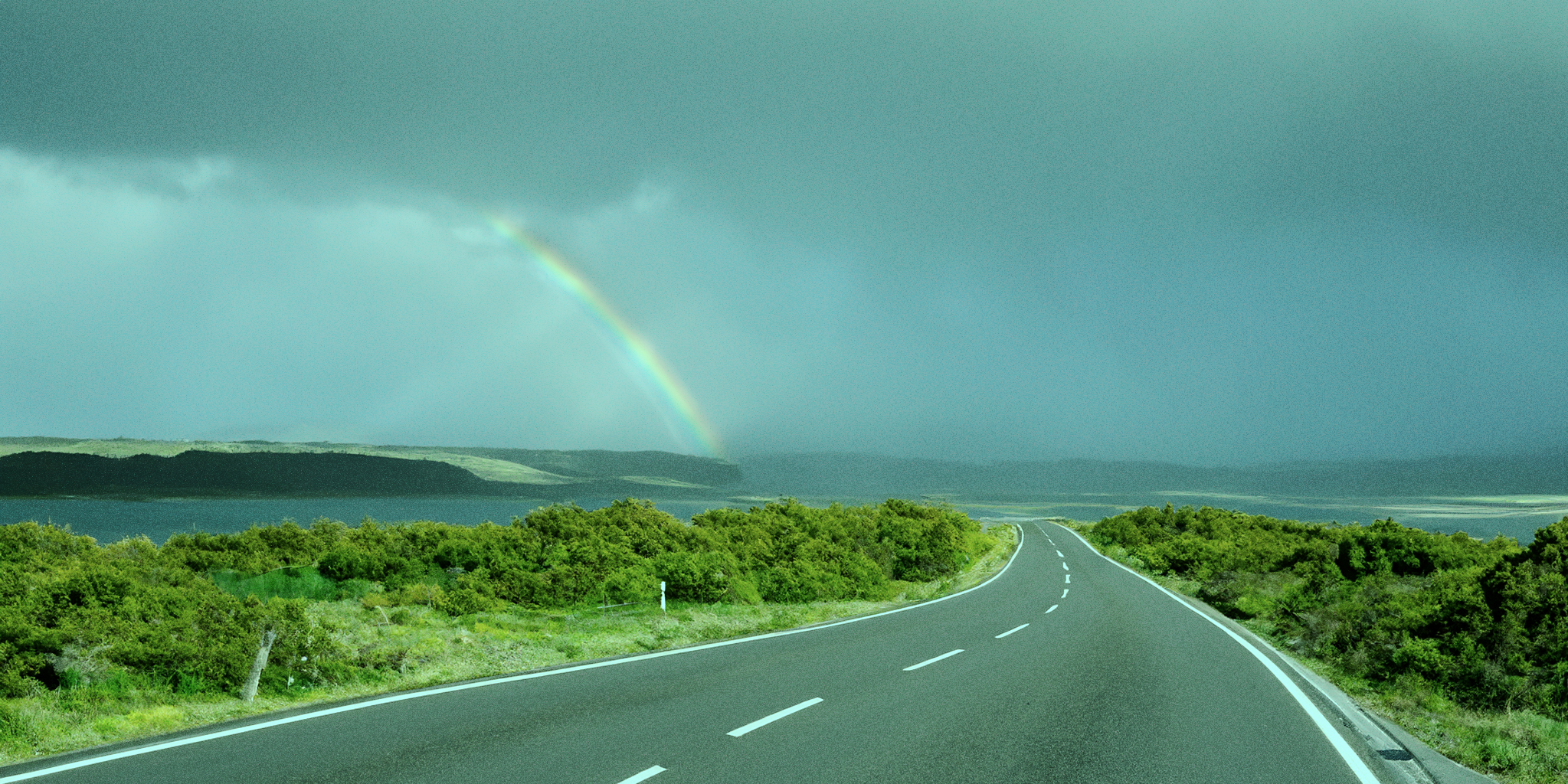 A winding road leads through lush green landscape under a stormy sky, with a faint rainbow appearing in the distance. The image symbolizes a journey with both challenges and hope ahead of a PMDD diagnosis.