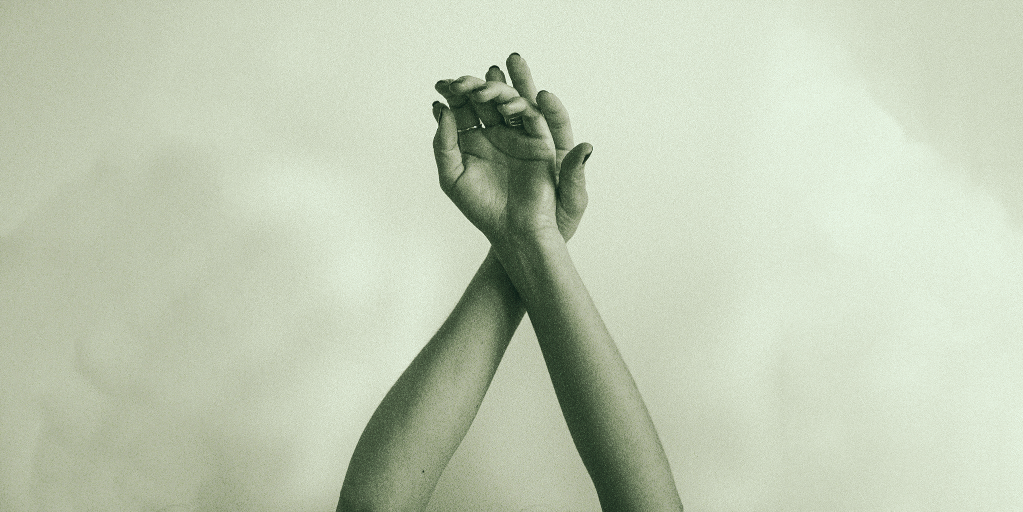 A pair of hands crossed at the wrists with fingers delicately intertwined. The image has a soft, monochromatic filter, giving it a calming, minimalistic aesthetic.