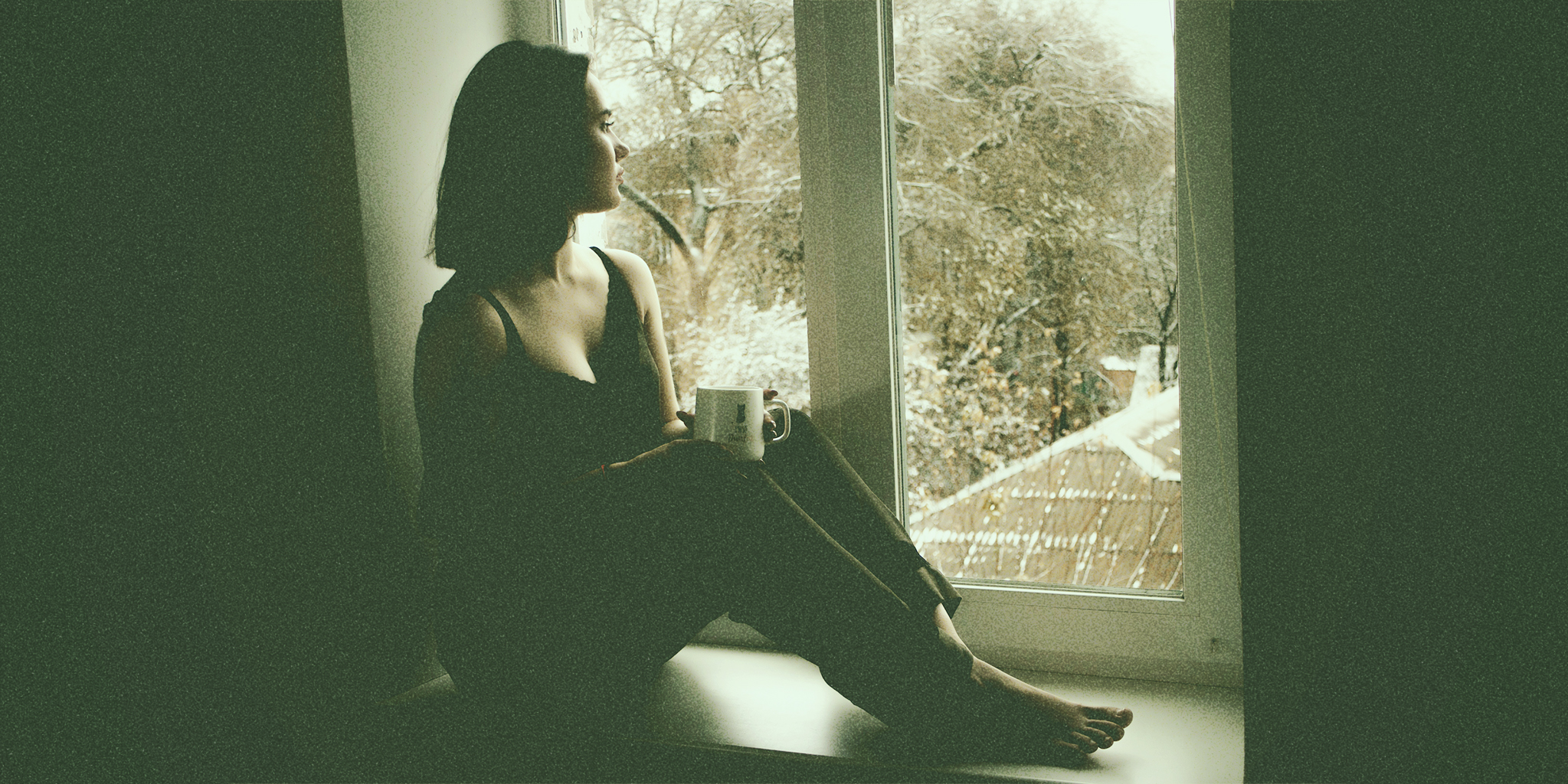 A woman sitting by a large window, holding a cup, gazing outside at a snowy scene. The soft natural light casts a subtle glow on her as she sits in a contemplative mood, framed by the outdoor view of snow-covered trees and rooftops.