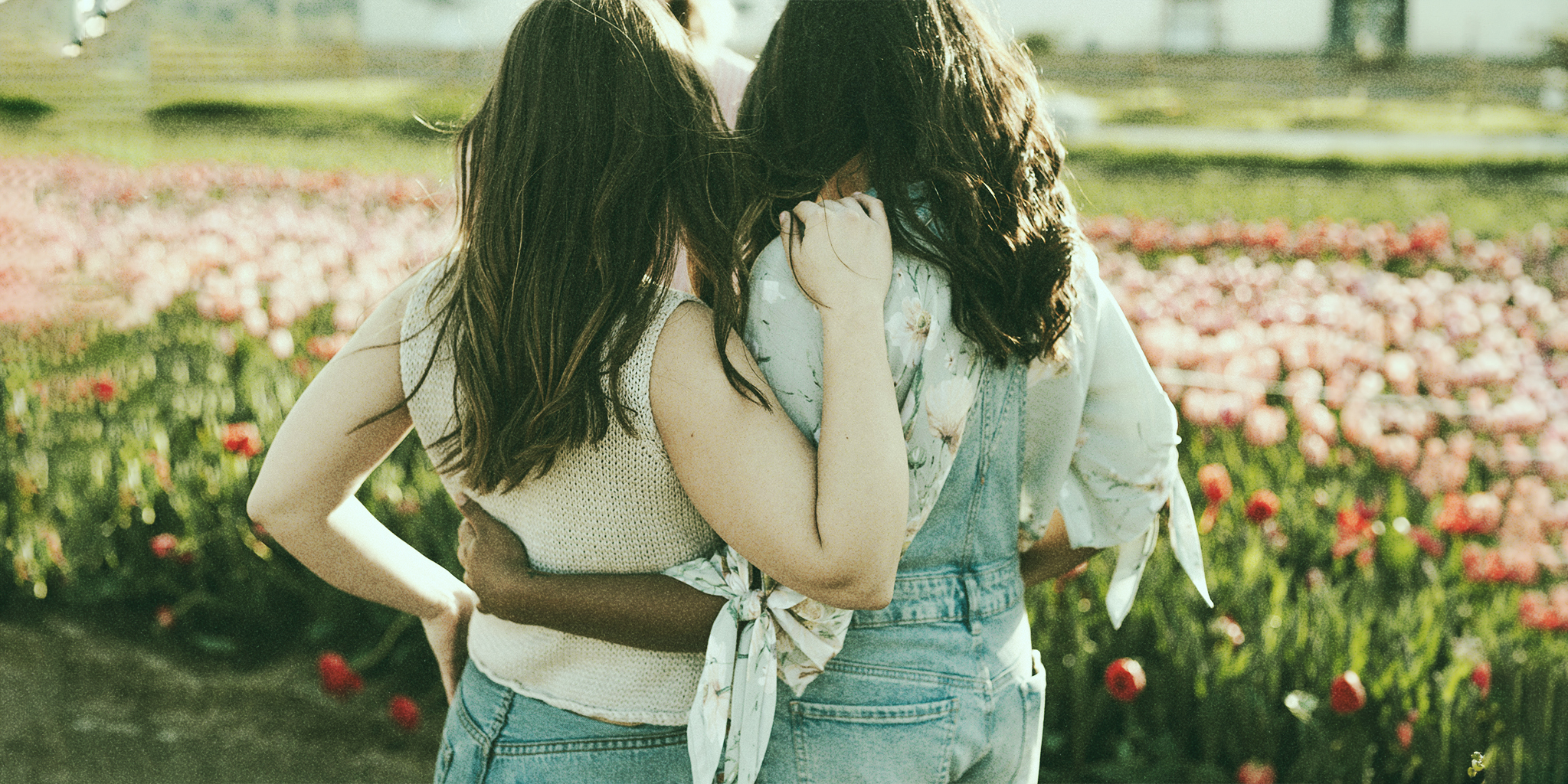 Two individuals standing closely with arms around each other, showing support and comfort, surrounded by blooming flowers.