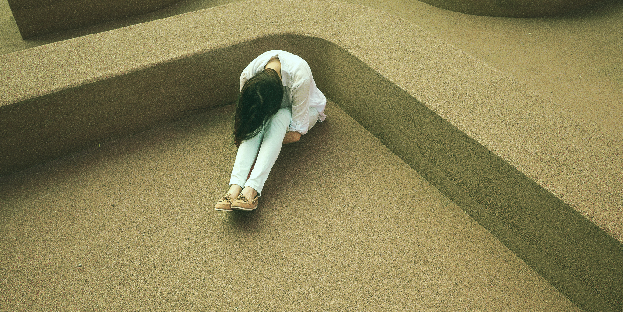 A woman sitting on the ground with her head lowered, hugging her knees. She is in an outdoor space with curved, textured concrete structures creating an abstract and isolated atmosphere.