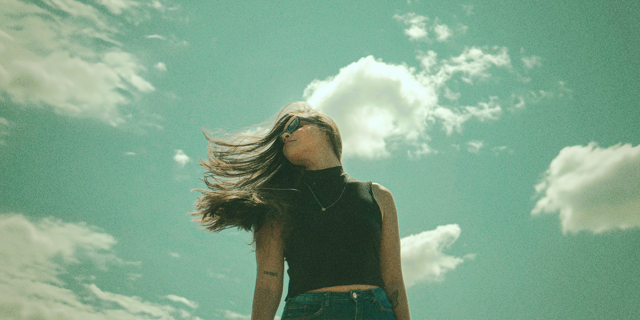 A young woman with long, flowing hair standing confidently against a bright sky with scattered clouds, symbolizing the journey of PCOS hair regrowth and renewal.