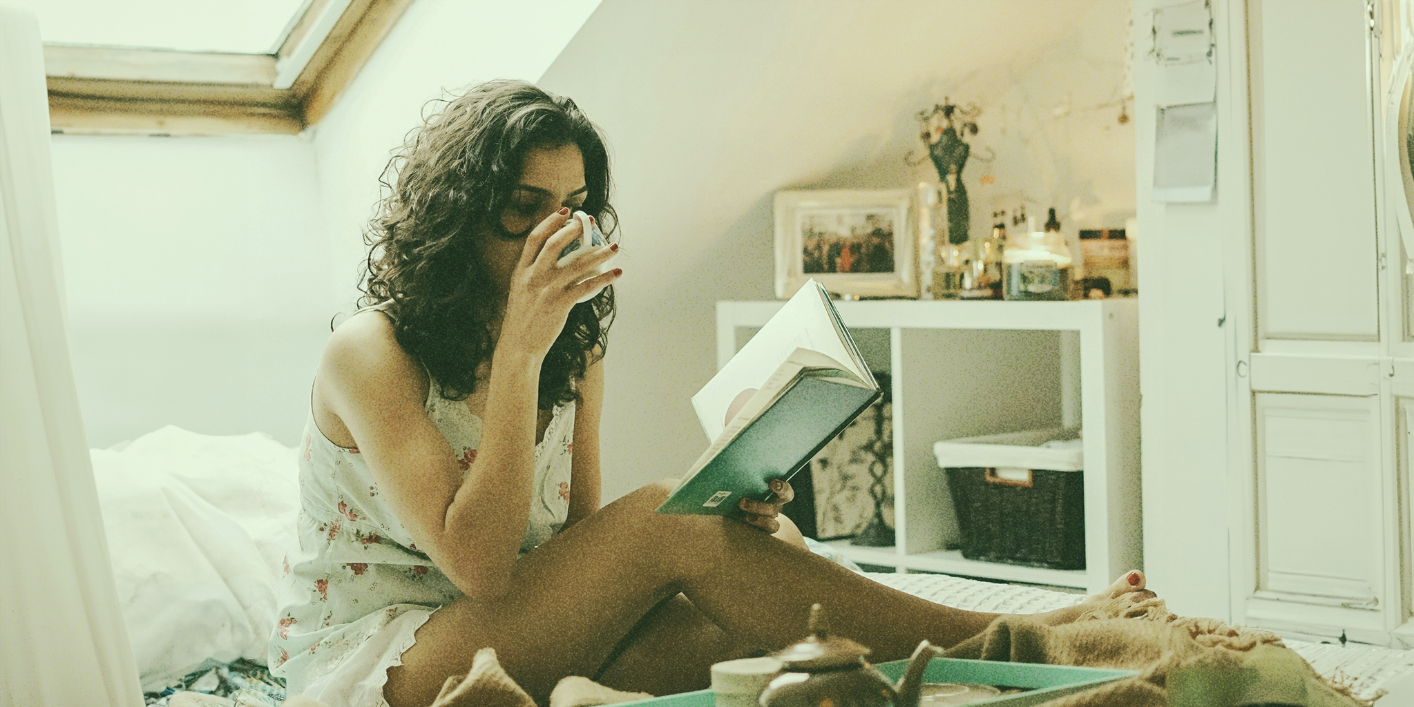 A woman in a floral dress sitting in a cozy, softly lit room, holding a cup of tea in one hand and reading a book with the other. The room features a skylight, shelves with personal items, and a relaxed ambiance.