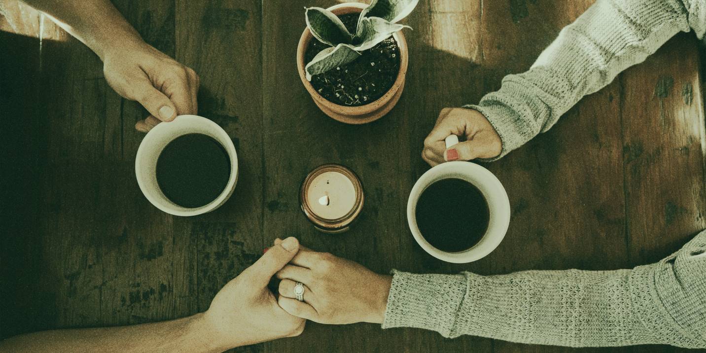 Two people holding hands over a wooden table, each holding a cup of coffee, with a potted plant and a lit candle in the center of the table.