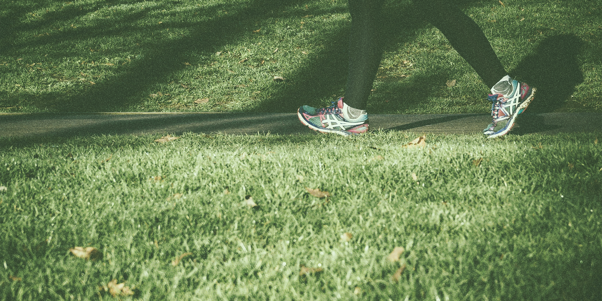 A person walking on a paved path in a grassy park, wearing colorful running shoes and black leggings. The sunlight creates dappled shadows across the grass. The image shows one of the answers to the question: "How to decrease progesterone?"