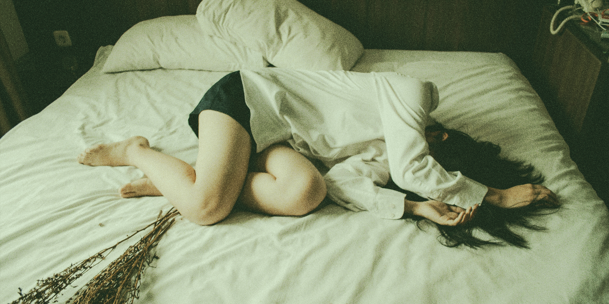A woman lying curled up on a bed, wearing a white shirt and black shorts, her face partially obscured. Dried flowers rest on the bed beside her, and the room has a dim, moody atmosphere. It shows the physical and emotional weight of hormonal changes: coping with pimples during ovulation.